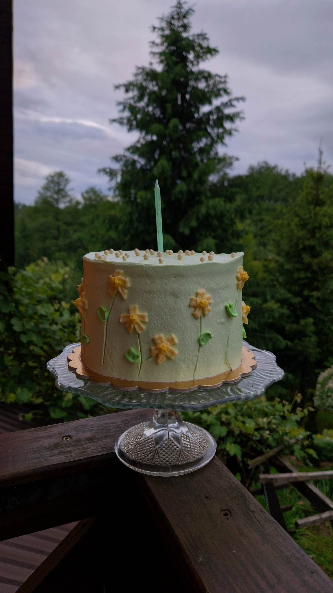 Tort urodzinowy z kremem w pastelowych odcieniach zieleni, ozdobiony kremowymi żółtymi kwiatami i zielonymi listkami, pojedyncza świeczka, na szklanym paterze, w tle zieleń drzew i pochmurne niebo.