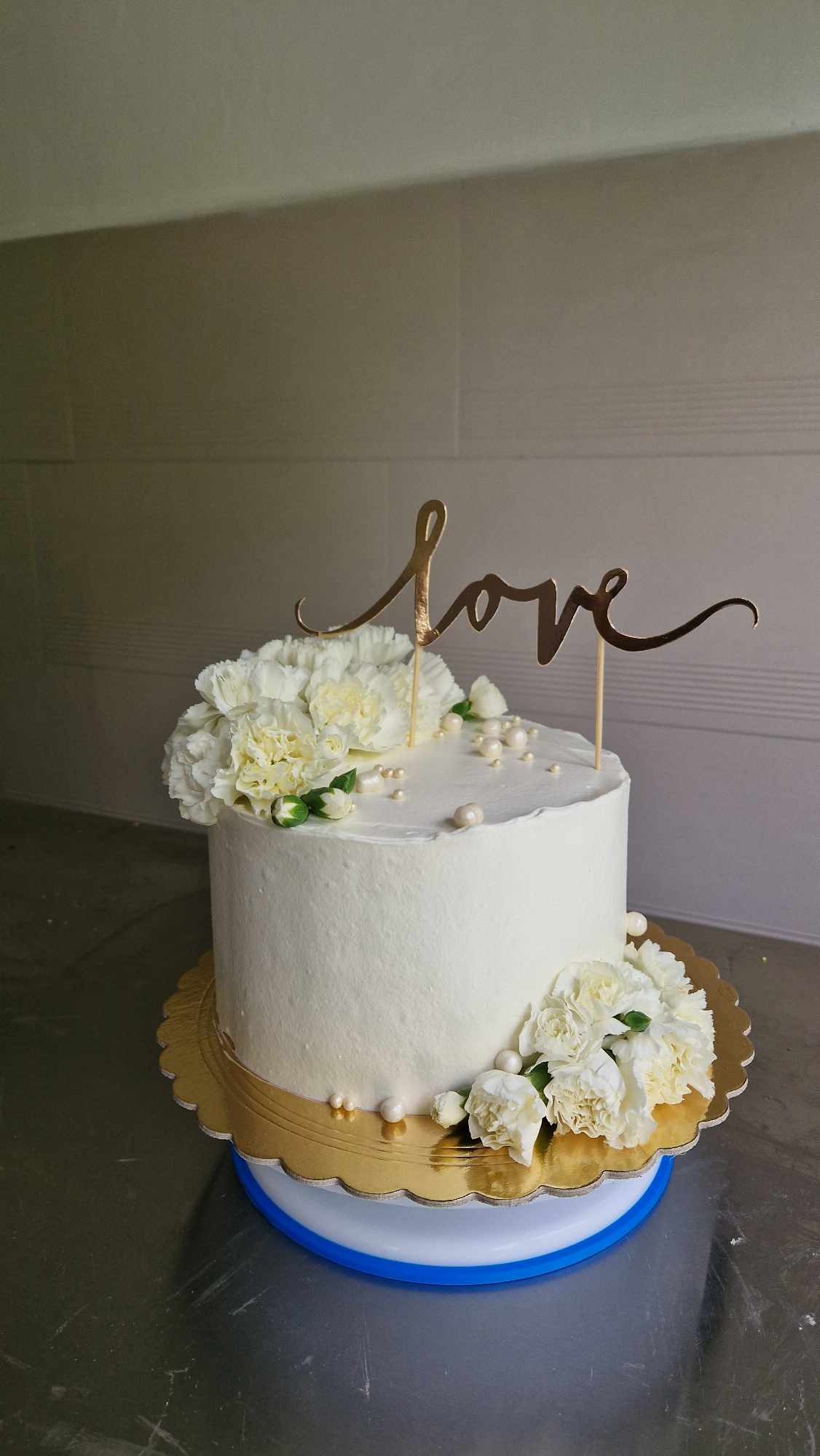 Tort weselny otynkowany białą masą, udekorowany białymi goździkami, perłowymi koralikami i złotym napisem 'love', umieszczony na złotej podkładce z niebieskim obrzeżem.