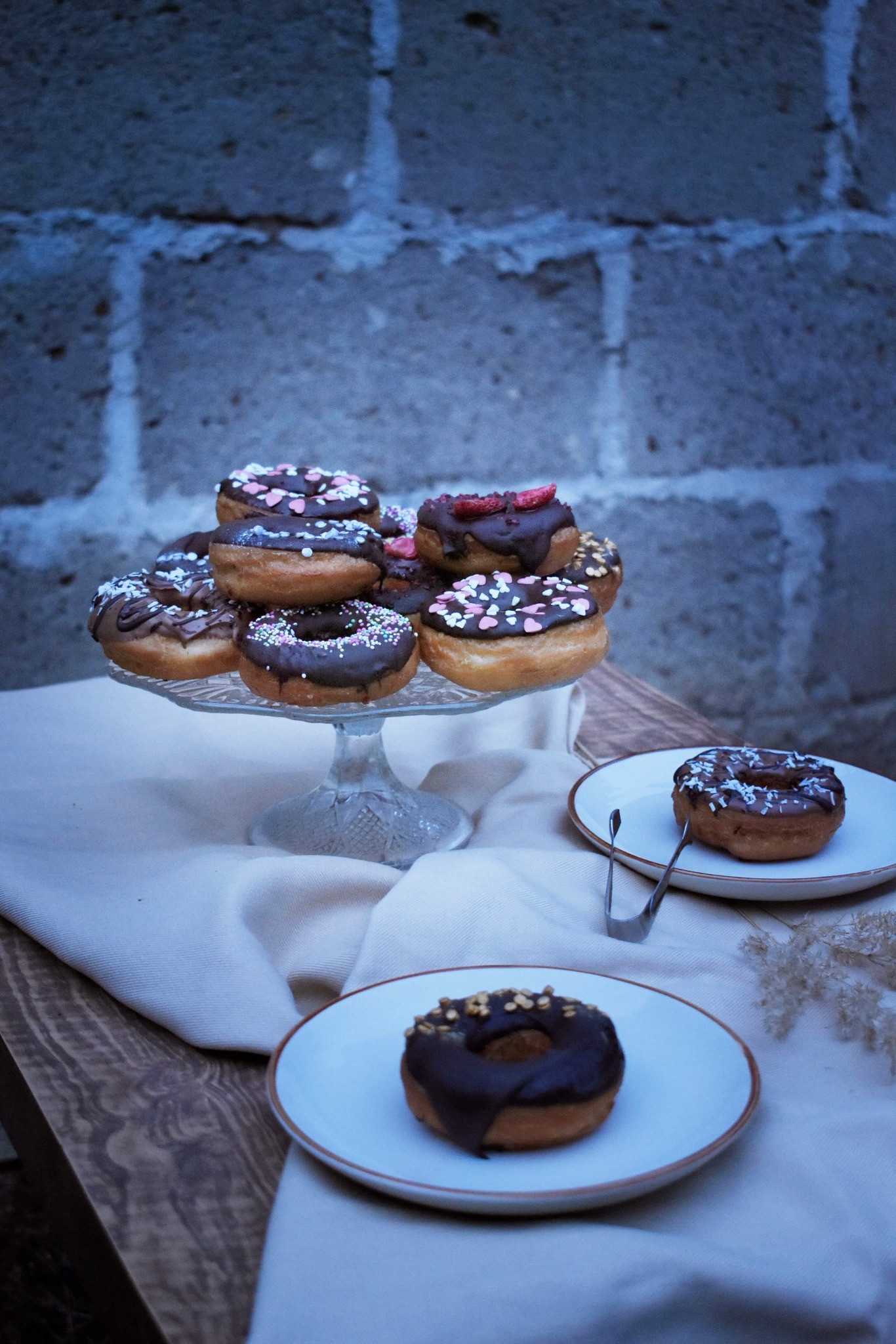 Stos pączków z czekoladową polewą i posypką na szklanym stojaku, dwa pączki na talerzykach obok, szczypce do ciasta, tło z cegieł.