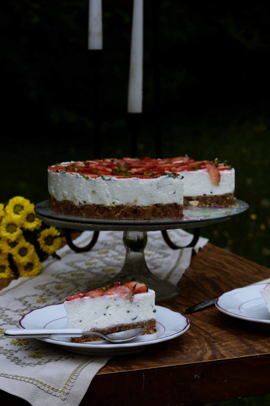Kawałek tortu truskawkowego z miętą na białym talerzyku z fioletową obwódką, obok srebrna łyżeczka, w tle cały tort na szklanej paterze, bukiet żółtych kwiatów i ciemne tło ogrodu.
