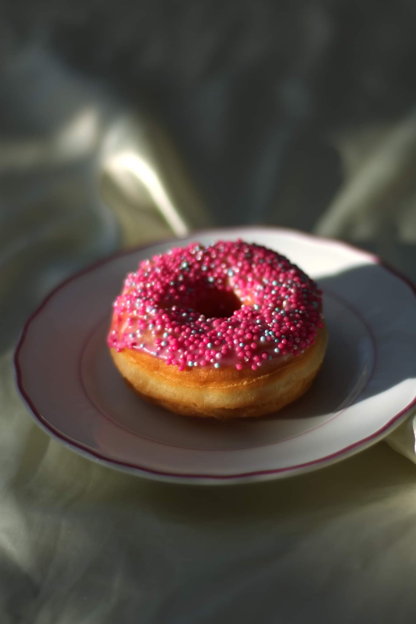 Pączek z różowym lukrem i posypką z drobnych, kolorowych kuleczek, leżący na białym talerzyku z delikatnym różowym obramowaniem, oświetlony miękkim światłem padającym z góry.