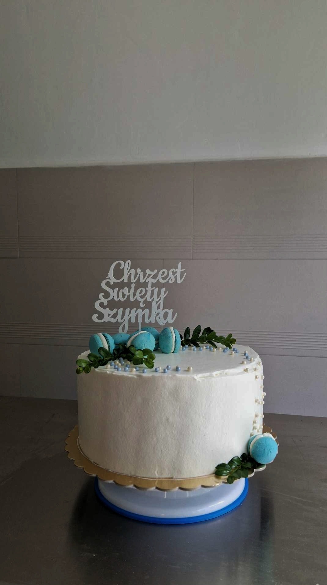 Tort na Chrzest Święty Szymka, udekorowany białym kremem, niebieskimi makaronikami, drobnymi perełkami i zielonymi listkami, z topperem z napisem.