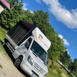 Biały bus transportowy z czarną plandeką na tle zieleni i błękitnego nieba z chmurami, zaparkowany na polnej drodze.