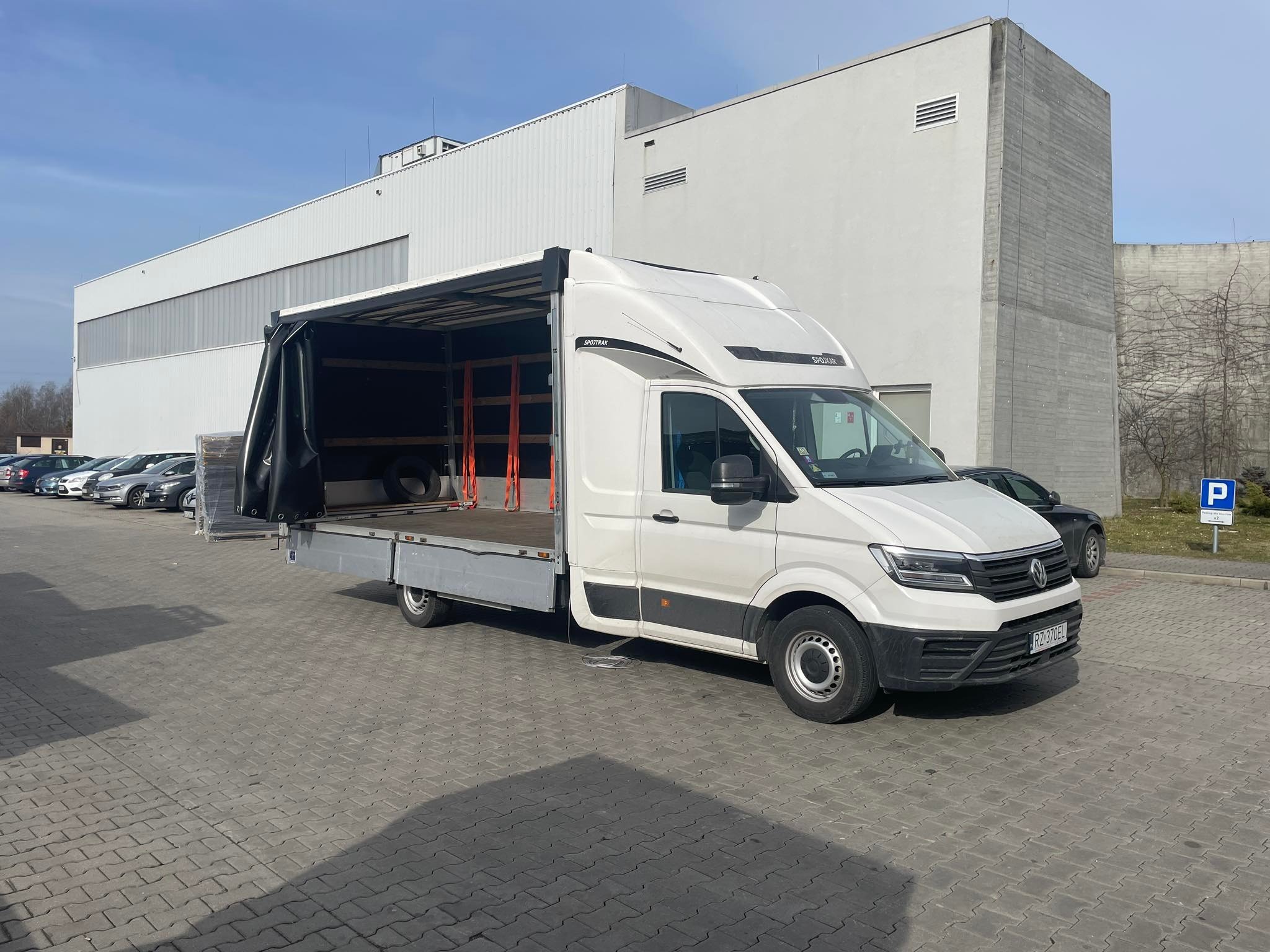 Biały samochód dostawczy z otwartą plandeką, zaparkowany na brukowanym placu przed budynkiem przemysłowym; wewnątrz widoczne zabezpieczenia ładunku i opona.