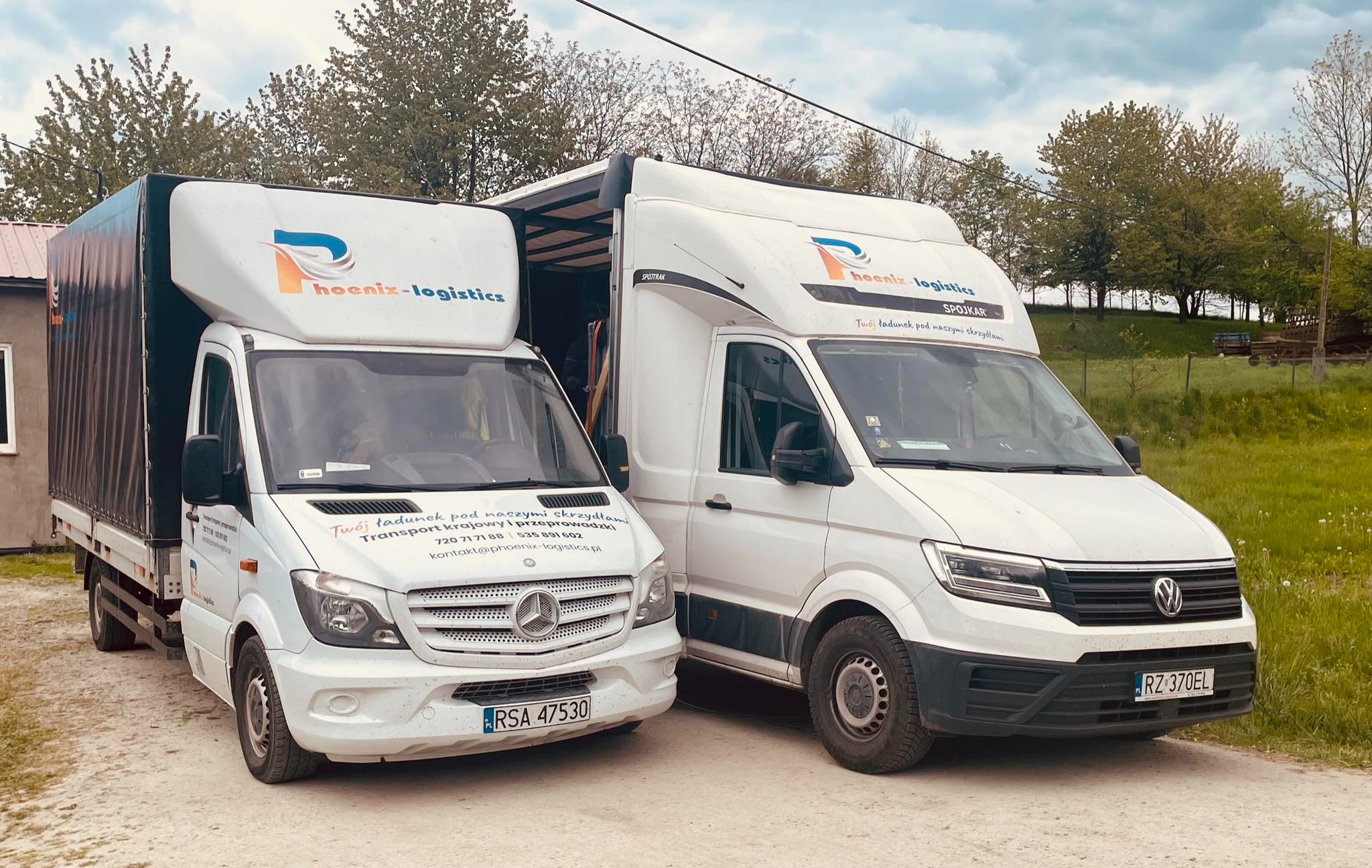 Dwa białe busy transportowe firmy Hoenix-Logistics zaparkowane obok siebie na polnej drodze, z widocznymi numerami rejestracyjnymi i logotypami firmy.