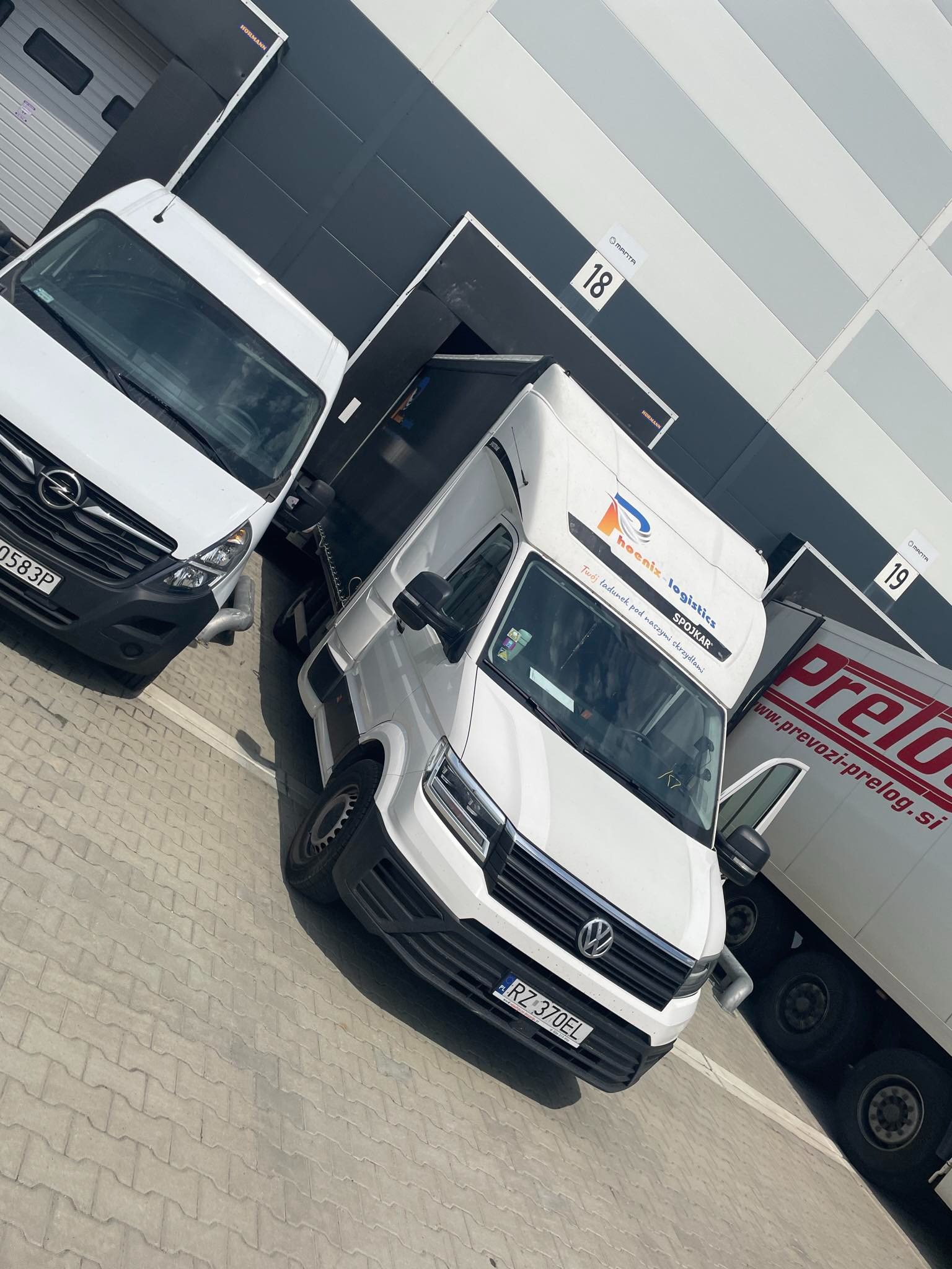 Trzy białe busy dostawcze zaparkowane przed rampami magazynowymi o numerach 18 i 19, w tym jeden z logo Phoenix Logistics Spojkar.