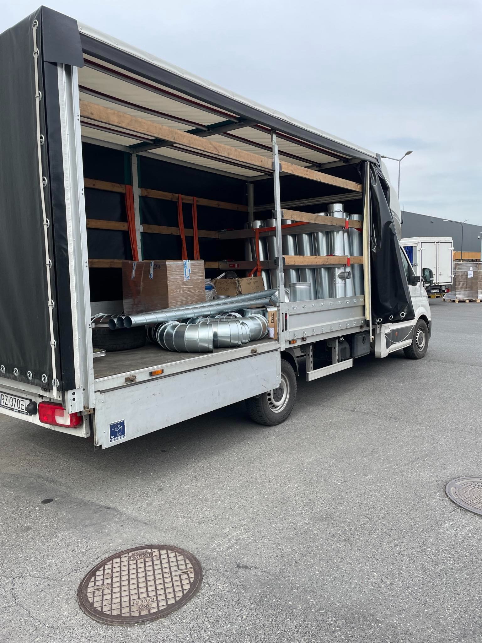 Otwarta paka samochodu dostawczego w Sanoku, załadowana metalowymi rurami wentylacyjnymi i kartonem, zabezpieczonymi pomarańczowymi pasami.