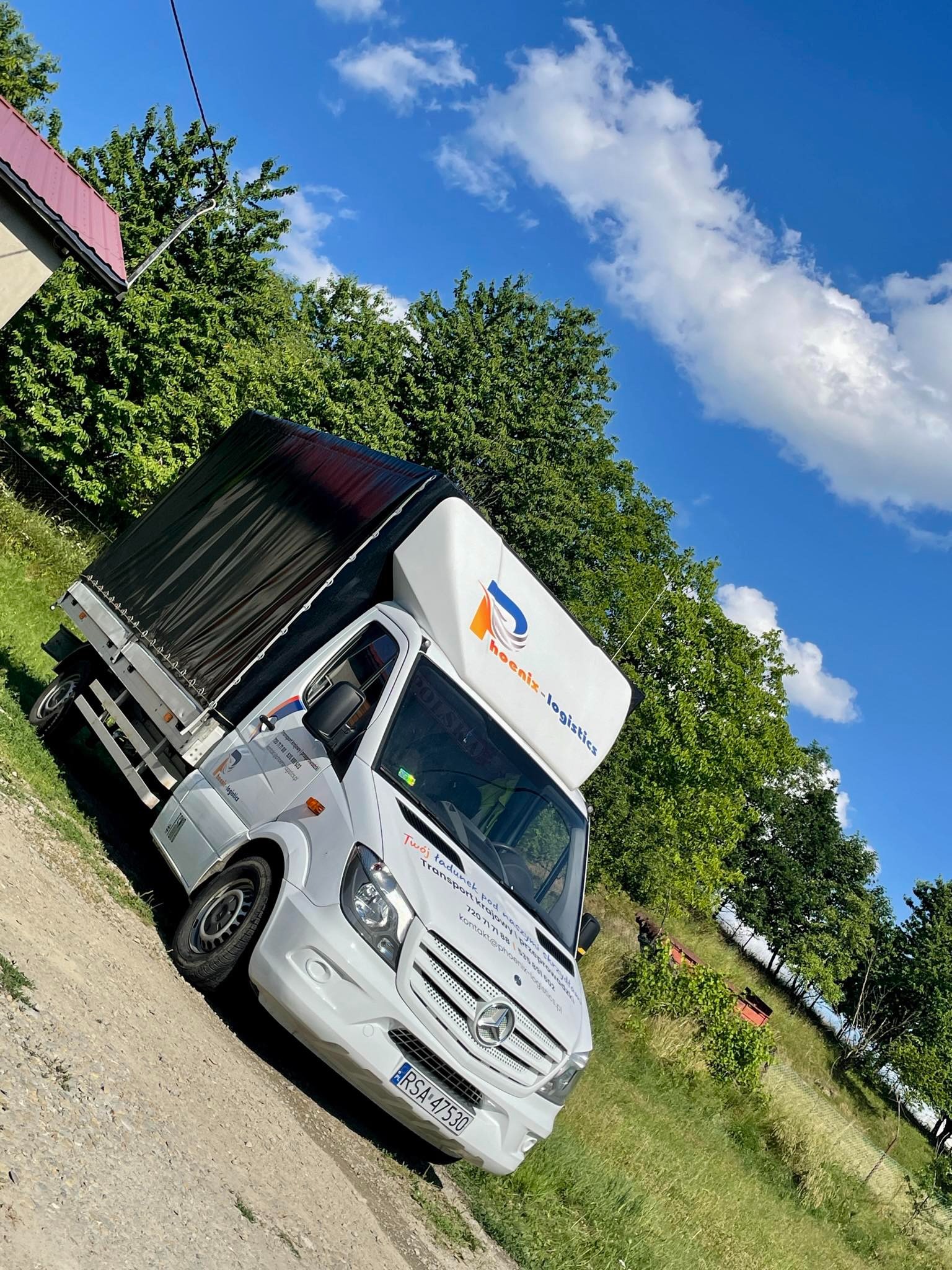 Biały bus transportowy z czarną plandeką na tle zieleni i błękitnego nieba z chmurami, zaparkowany na polnej drodze.