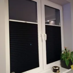 Ciemne, plisowane osłony okienne zamontowane w białym oknie PCV, z widocznymi dekoracjami na parapecie: świecznik, zegar i roślina doniczkowa.