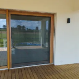 Zewnętrzna moskitiera w drewnianej ramie, zamontowana na tarasie z desek, widok na ogród w tle.