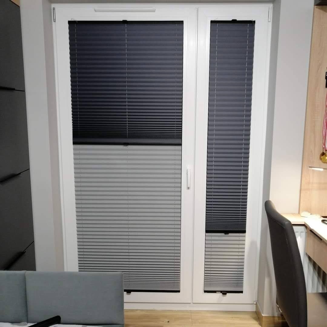 Dwuskrzydłowe okno z zamontowanymi plisowanymi osłonami okiennymi, częściowo zasłoniętymi, z widocznym mechanizmem sterującym.