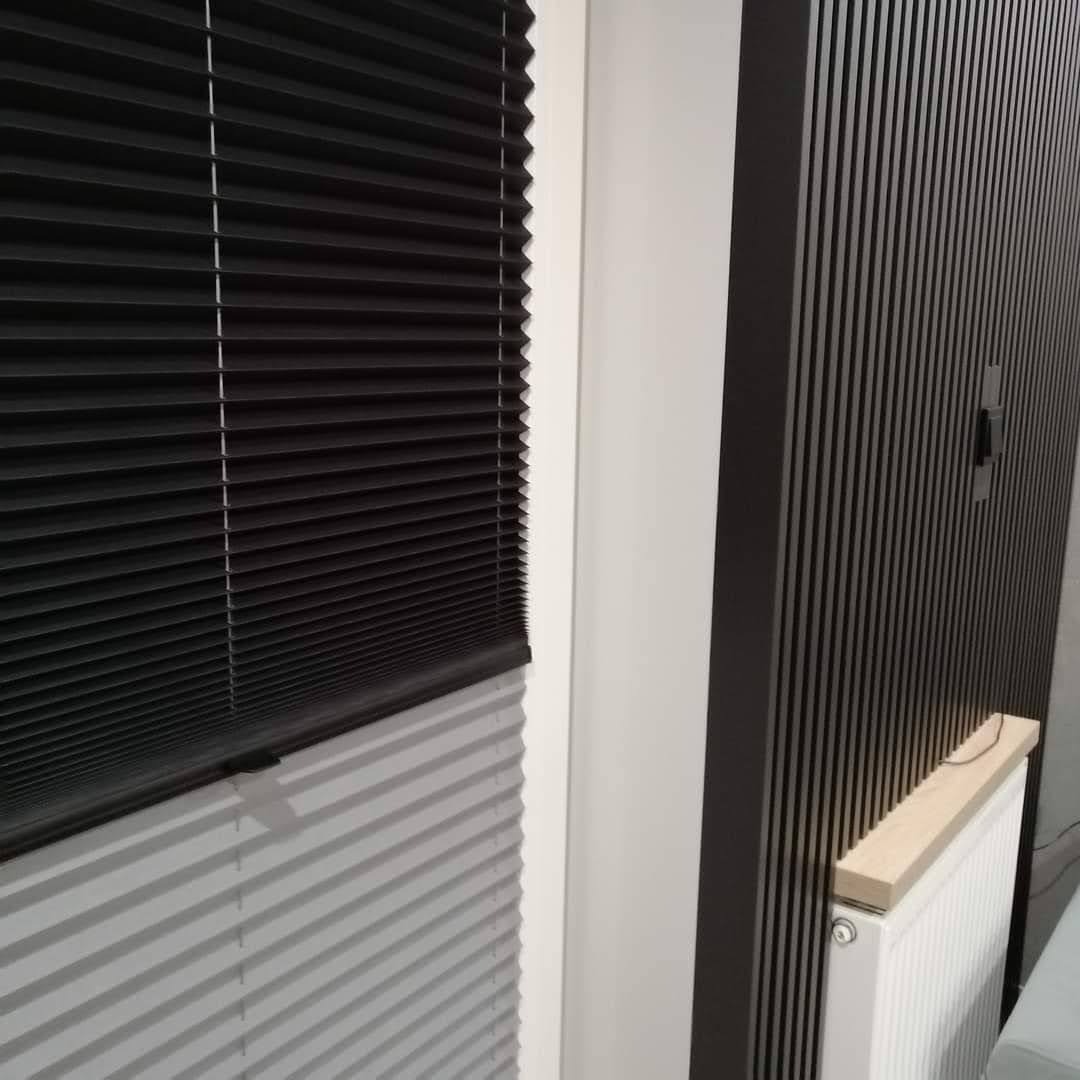 Częściowo opuszczona, czarna plisa okienna w nowoczesnym wnętrzu, widoczny fragment grzejnika z drewnianym elementem i pionowe lamele na ścianie.