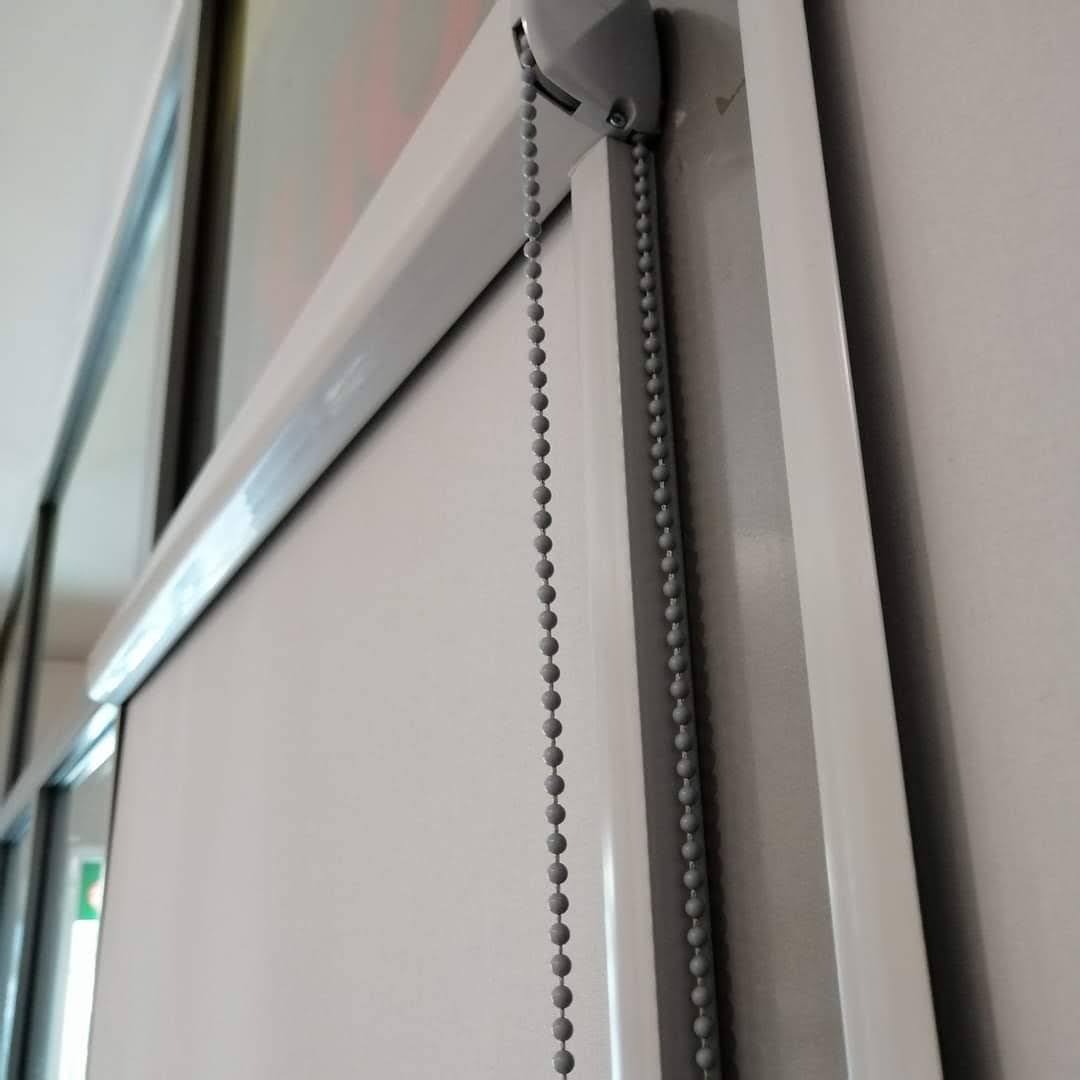 Zamknięta, biała roleta z łańcuszkiem kulkowym, widok z dołu na tle okna.