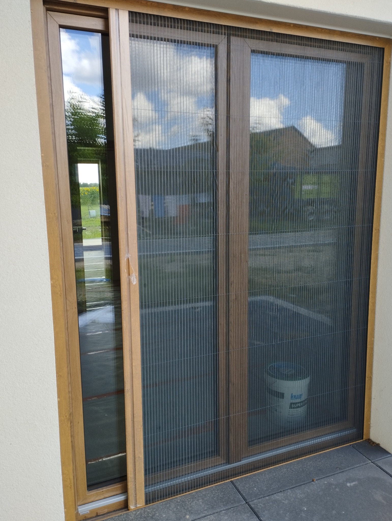 Zewnętrzna moskitiera plisowana w brązowej ramie na drzwiach tarasowych, widoczny krajobraz i wiadro z klejem Knauf za siatką.