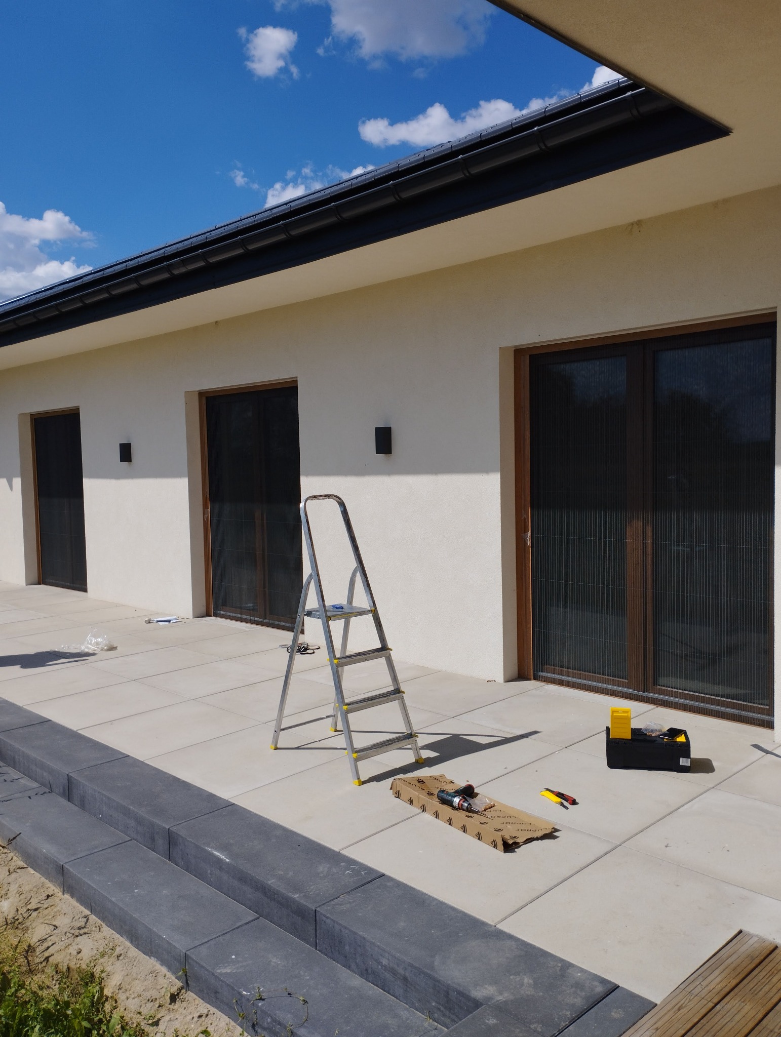 Montaż rolet plisowanych w oknach tarasowych nowoczesnego domu; widoczna drabina, narzędzia i skrzynka narzędziowa na jasnym tarasie.