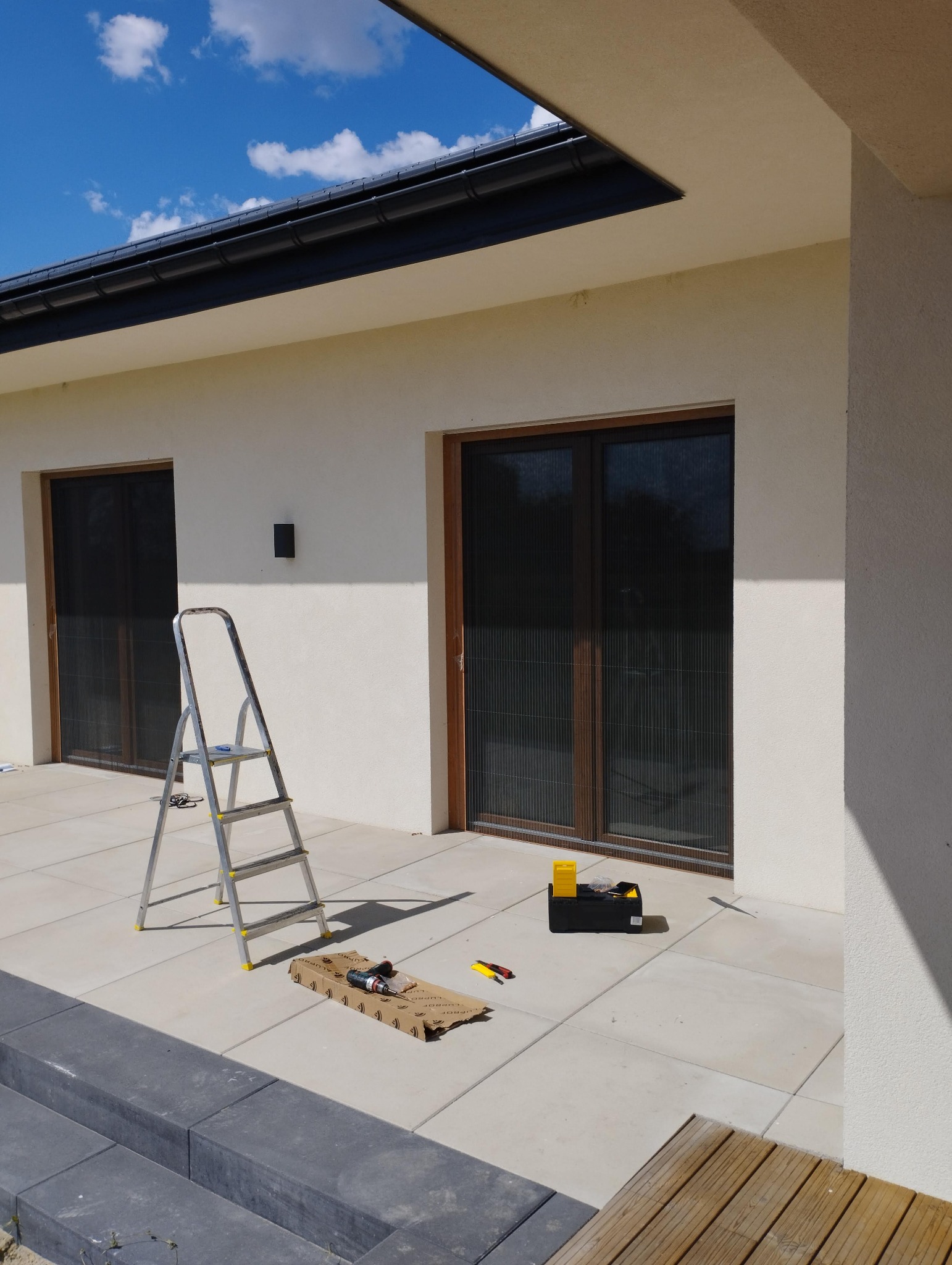 Montaż moskitier plisowanych w oknach tarasowych, widoczna drabina, skrzynka z narzędziami i wkrętarka na kartonie.