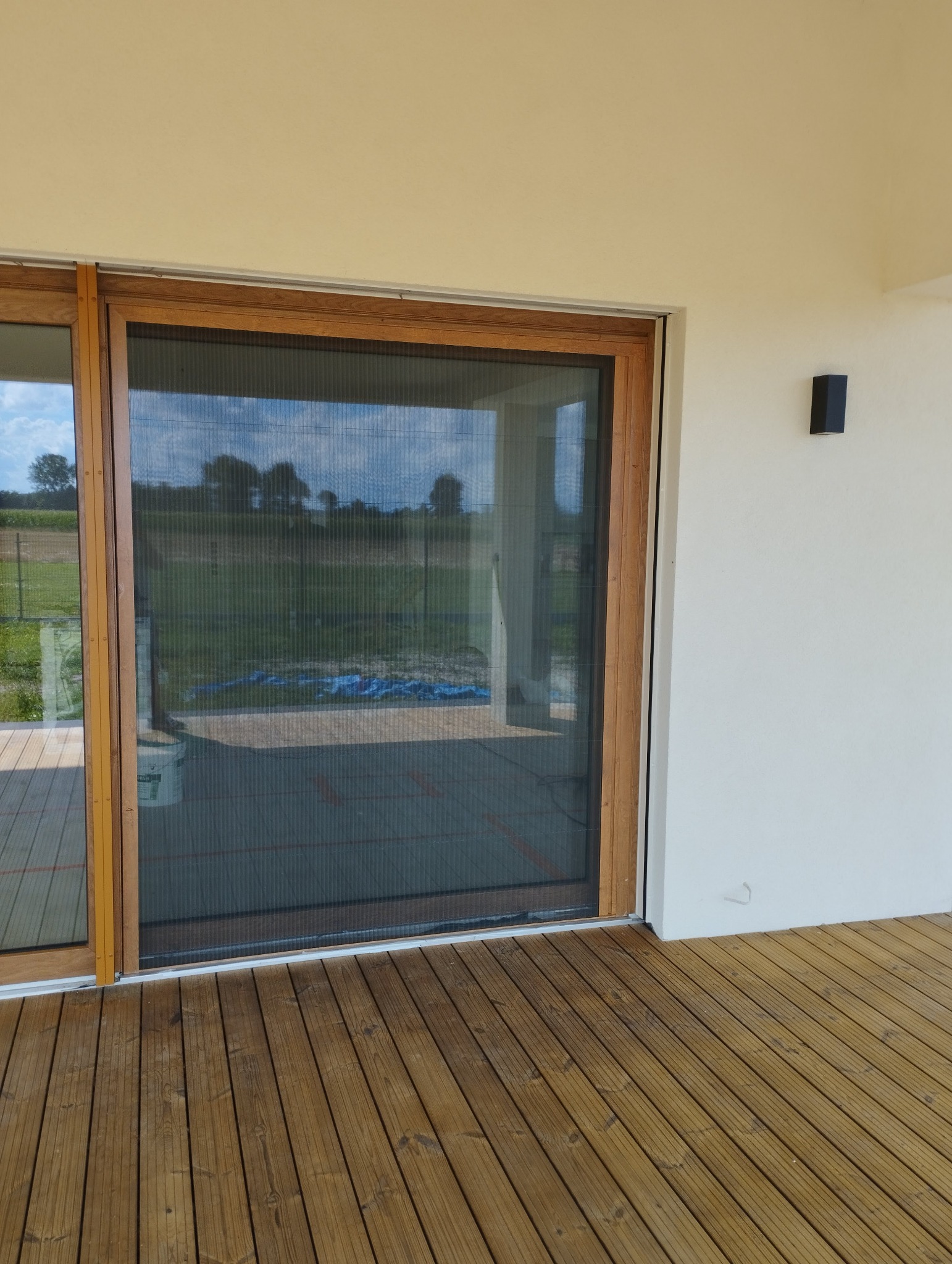 Zewnętrzna moskitiera w drewnianej ramie, zamontowana na tarasie z desek, widok na ogród w tle.
