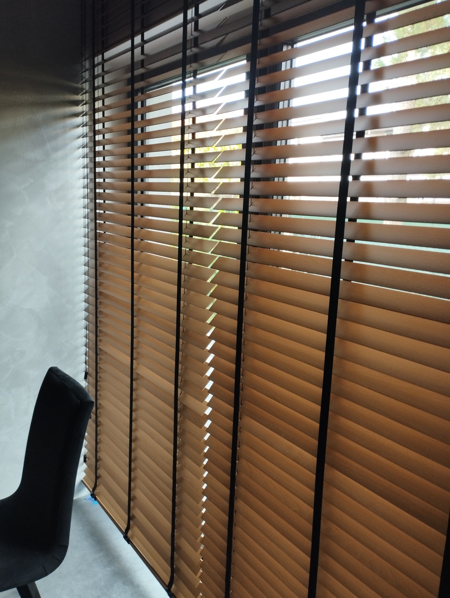 Zamknięte drewniane żaluzje poziome w odcieniach brązu, z widocznymi sznurkami i fragmentem czarnego krzesła w rogu zdjęcia.