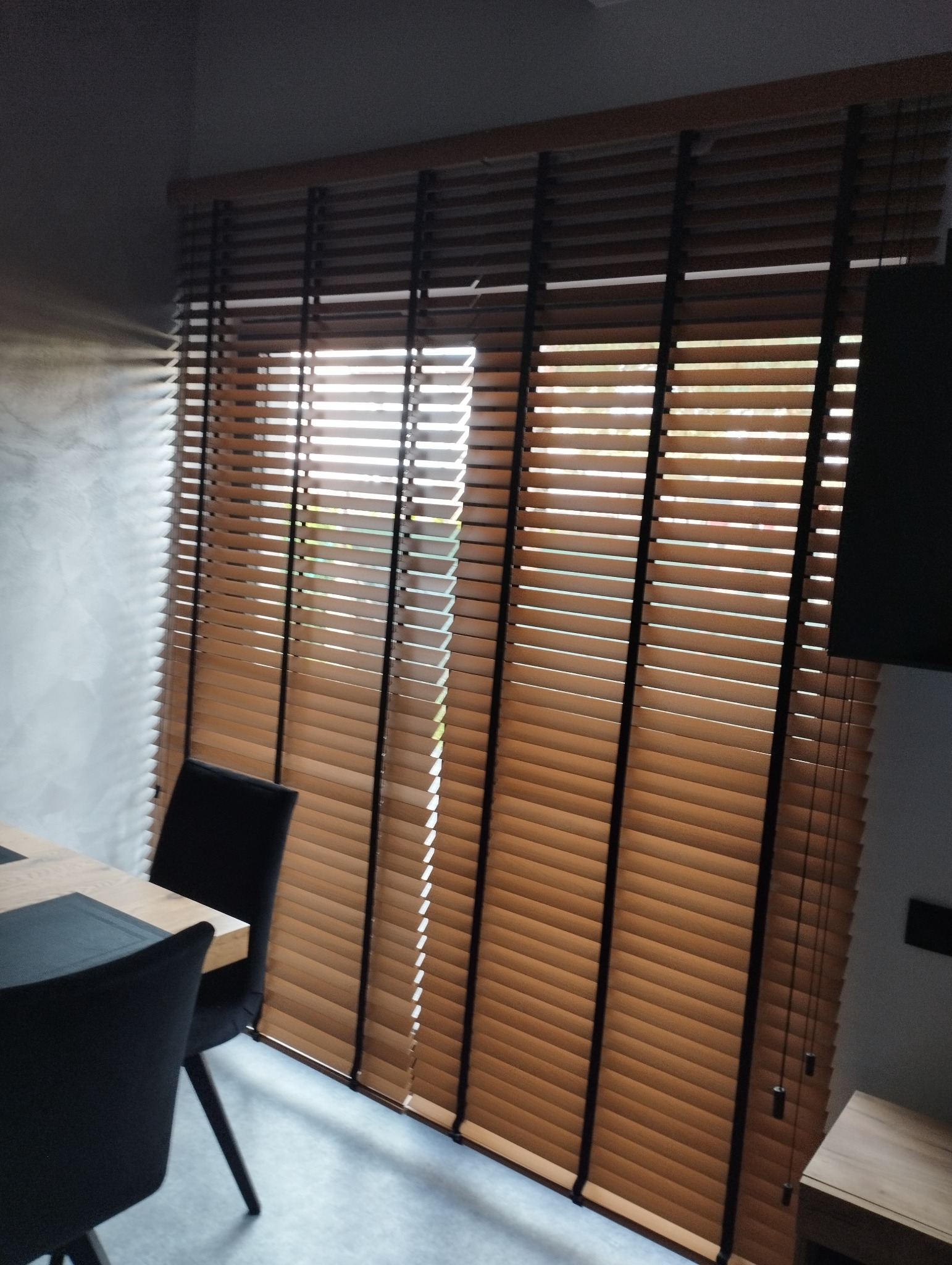 Drewniane żaluzje poziome w kolorze jasnego drewna, zamontowane w oknie, z widocznym fragmentem stołu i krzesła w minimalistycznym wnętrzu.