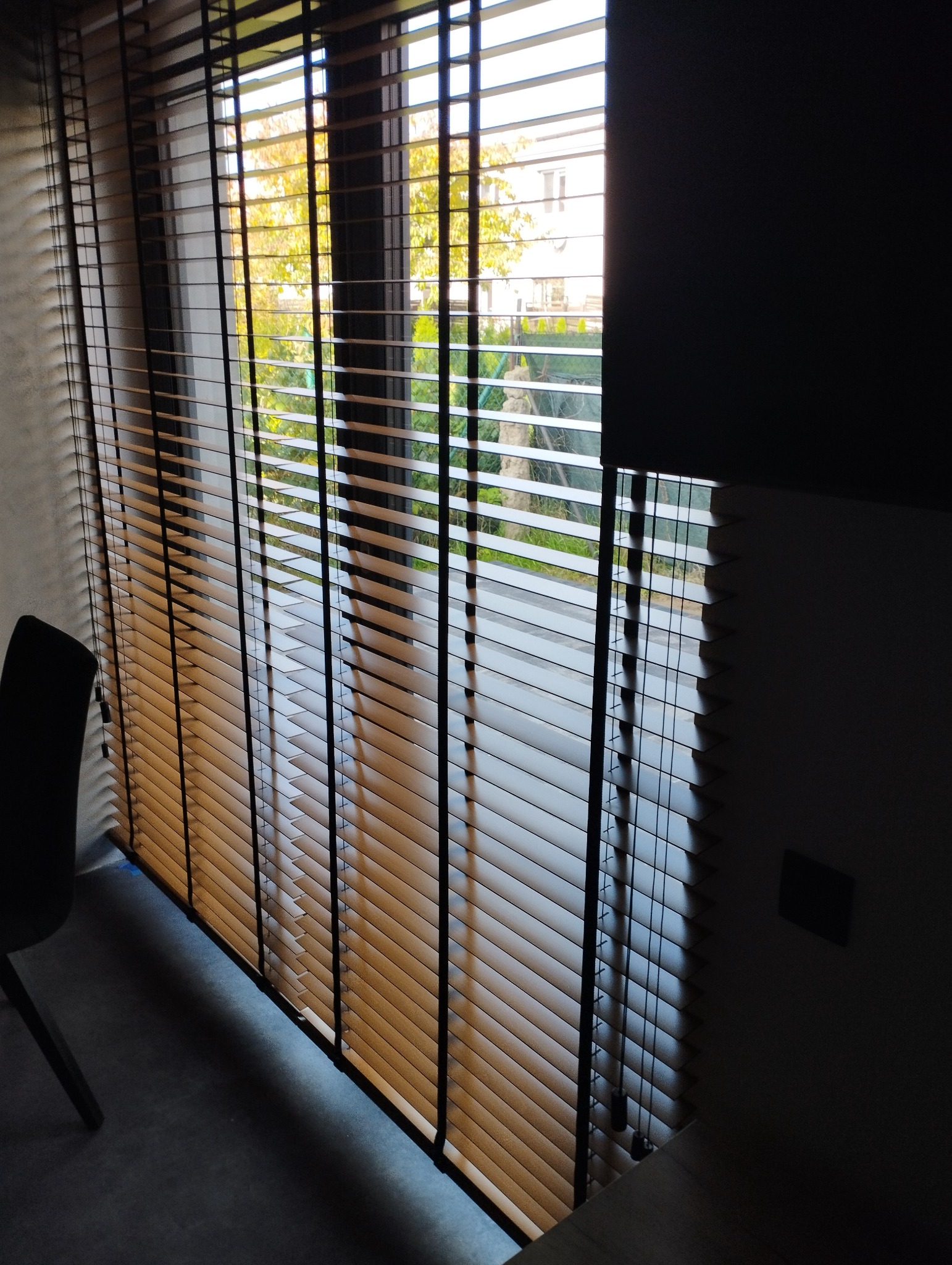 Brązowe żaluzje drewniane z czarnymi sznurkami, częściowo zasłonięte, wpadające światło słoneczne, widok na dom i zieleń na zewnątrz.