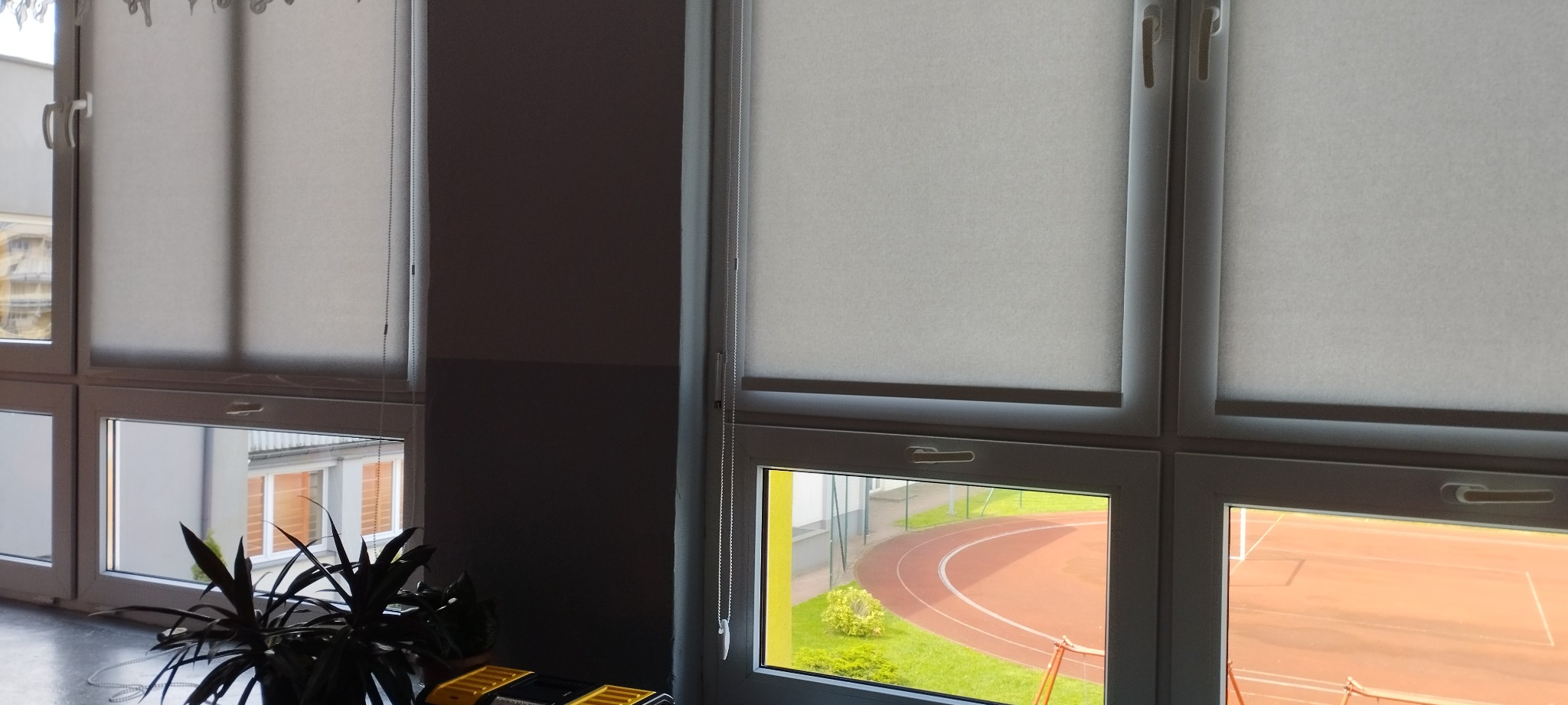 Zasłonięte okna z widokiem na bieżnię i kort tenisowy przez białe, półprzezroczyste rolety w kasecie z łańcuszkiem.