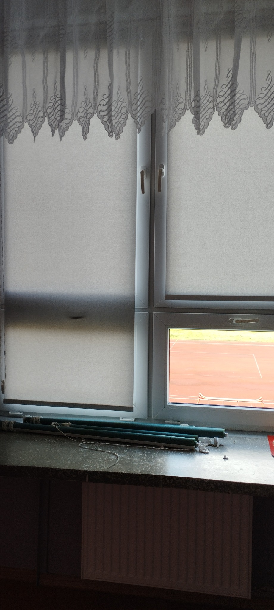 Okno z zamontowaną roletą dzień noc, widoczne zwinięte wałki rolety na parapecie, za oknem stadion lekkoatletyczny.