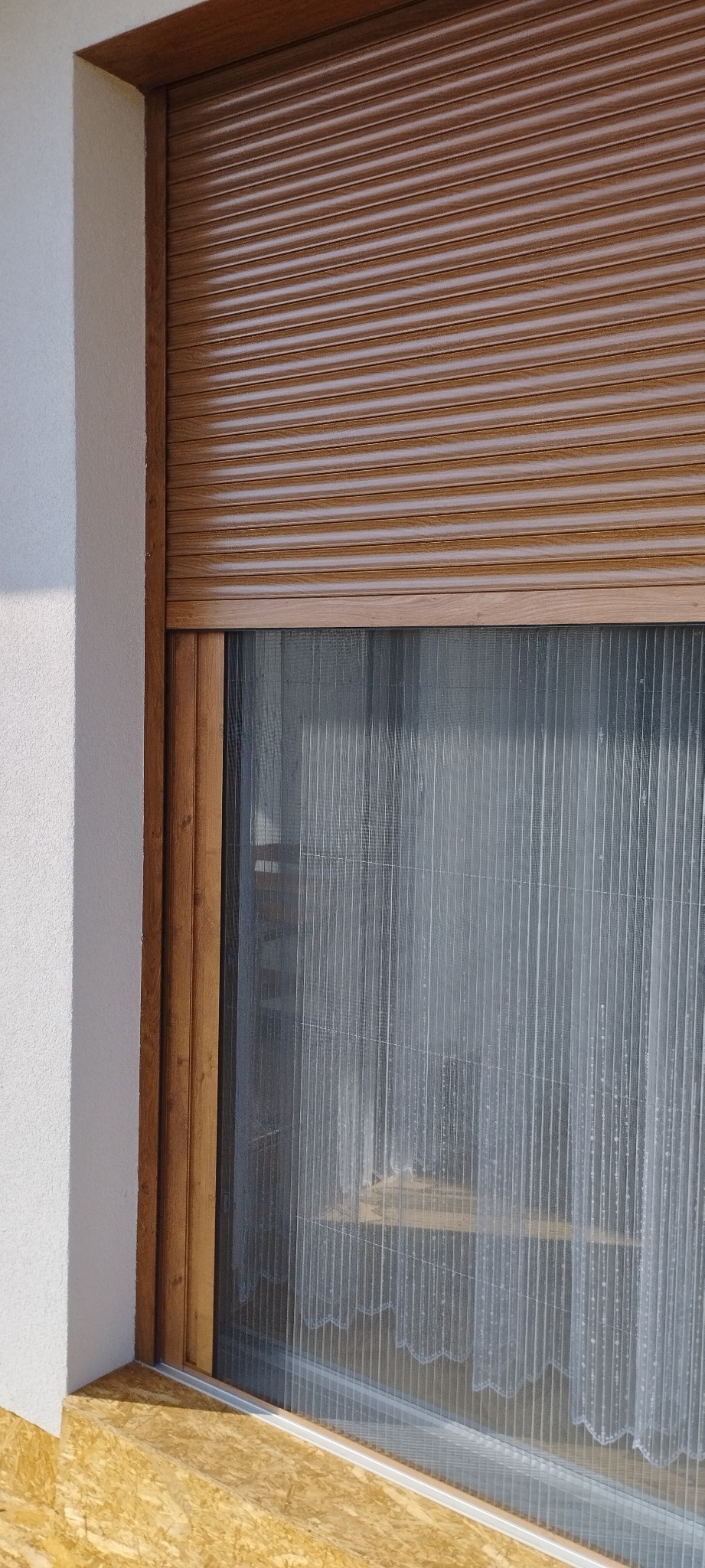 Zasłonięte okno z brązową roletą zewnętrzną, widoczna moskitiera i firanka wewnątrz.