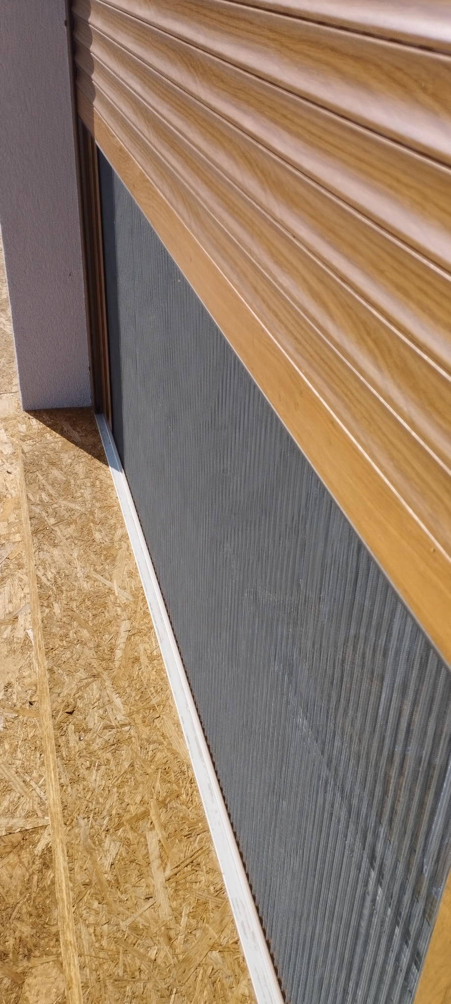Fragment zamkniętej, drewnopodobnej rolety zewnętrznej z widoczną fakturą lamel i szarą powierzchnią pod spodem, zamontowanej na surowej płycie OSB.