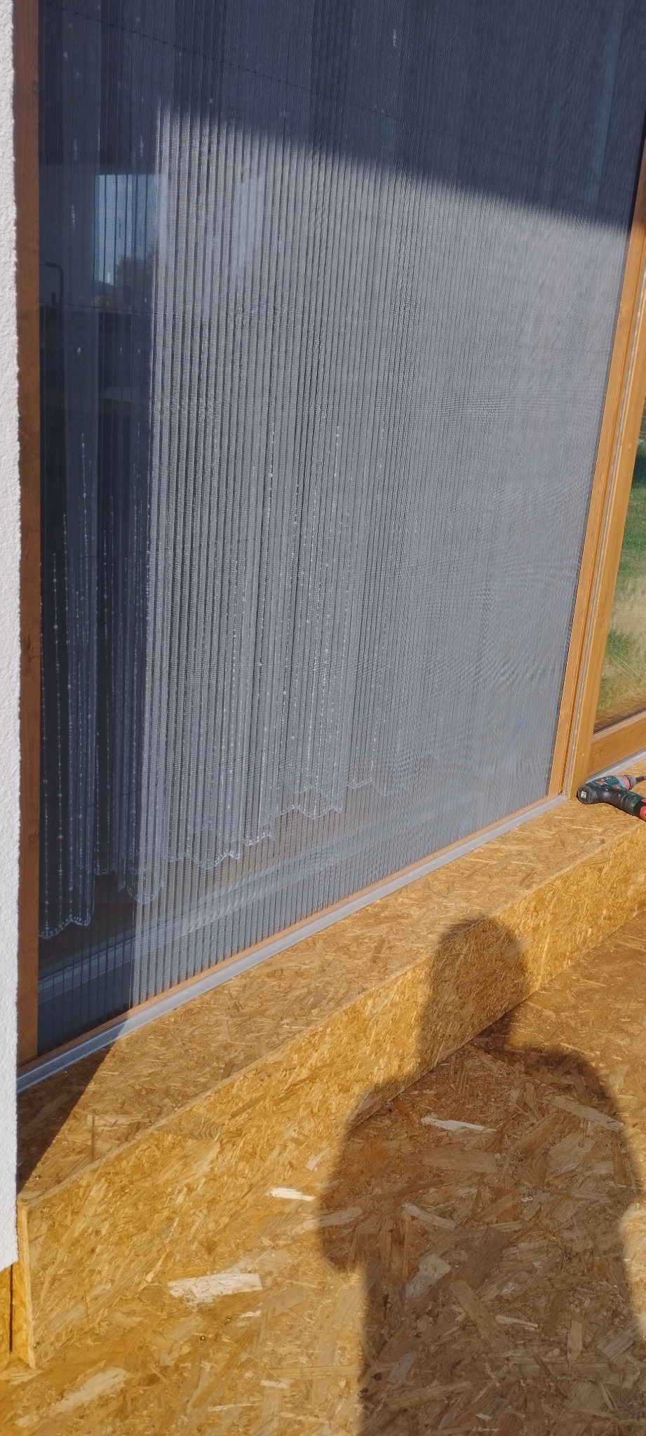 Szara, plisowana przesłona okienna w drewnianej ramie, widoczna od wewnątrz, z shadow osoby montującej na drewnianym parapecie.