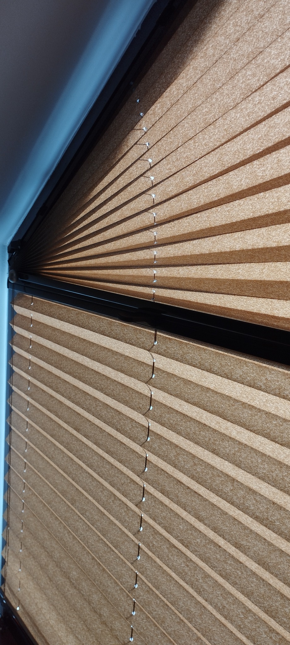 Beżowe rolety plisowane z czarną kasetą, widoczne sznurki regulujące, zamontowane w oknie z niebieskawym światłem.