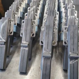 Rząd metalowych, spawanych elementów konstrukcyjnych, prawdopodobnie fragmenty balustrady lub poręczy, ułożonych na metalowym stole warsztatowym, widoczne ślady spawania i obróbki.