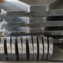 Stos metalowych elementów o trapezoidalnym kształcie, z widocznymi spawami i otworami montażowymi, ułożonych jeden na drugim w warsztacie.