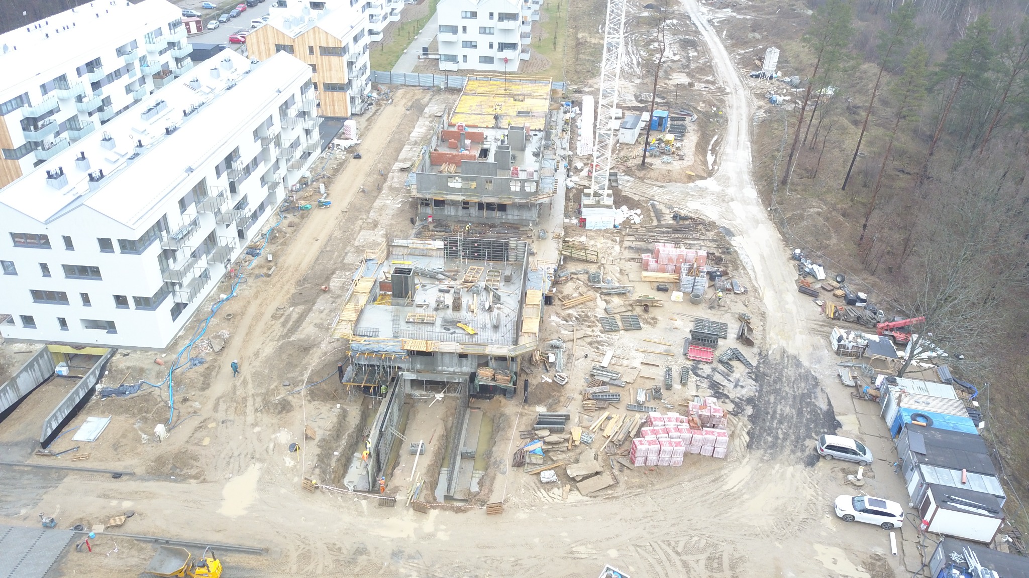 Widok z lotu ptaka na plac budowy w Olsztynie z trzema budynkami w stanie surowym, z widocznymi fundamentami, ścianami i elementami konstrukcji żelbetowej, obok stoją gotowe budynki mieszkalne.