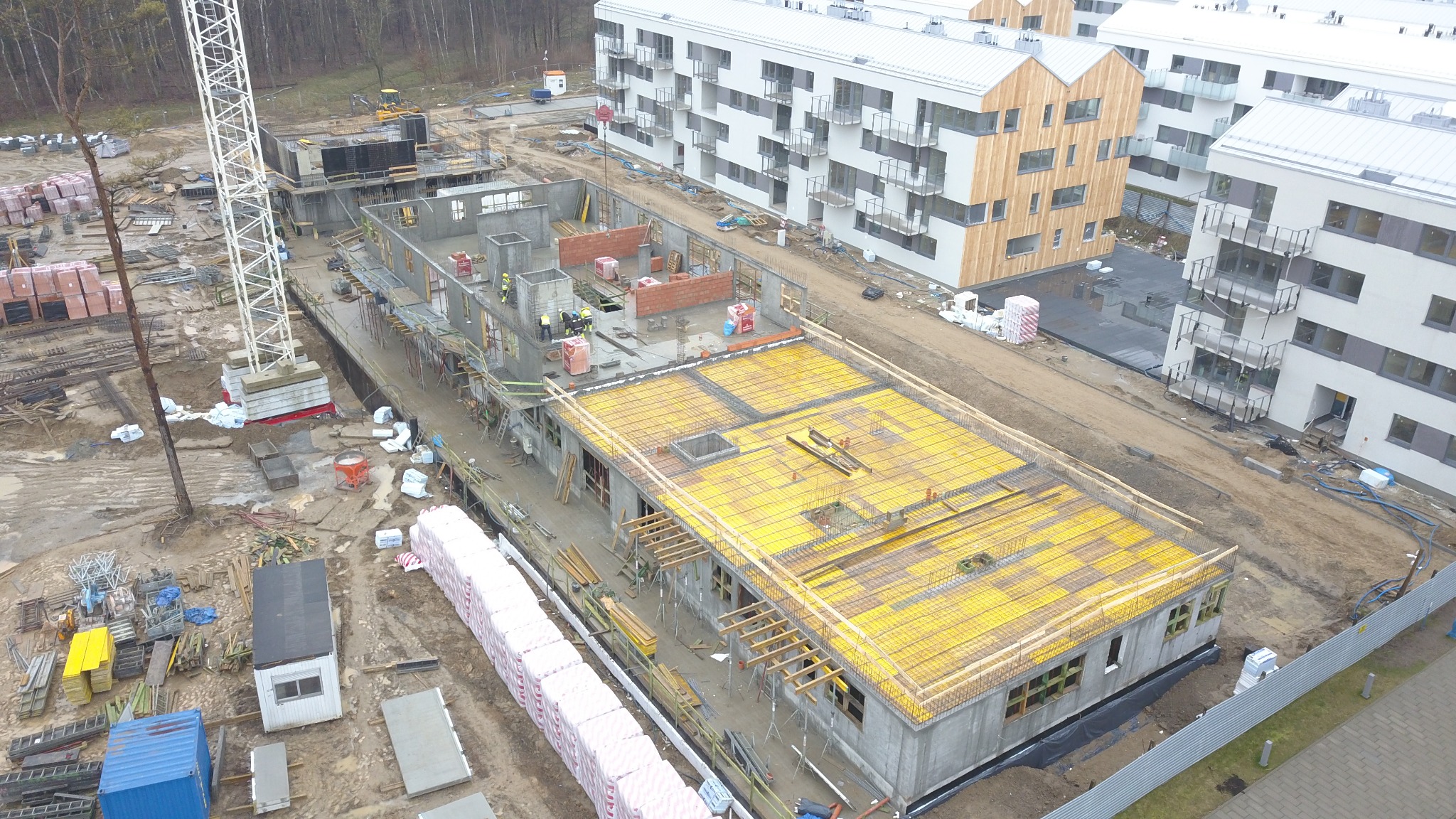 Budowa budynku żelbetowego: widok z góry na plac budowy z częściowo wzniesionymi ścianami, żółtym szalunkiem stropowym, pracownikami w kaskach oraz stosem czerwonej cegły i białych bloczków.