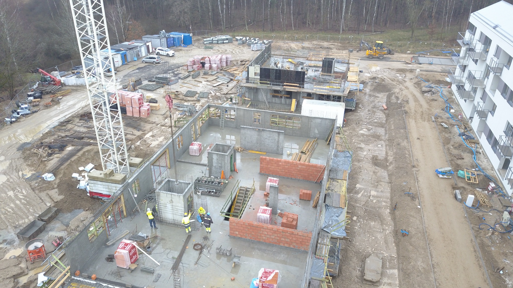 Budowa budynku wielorodzinnego w trakcie wznoszenia ścian, widok z góry na plac budowy z dźwigiem, materiałami budowlanymi i pracownikami w żółtych kamizelkach.