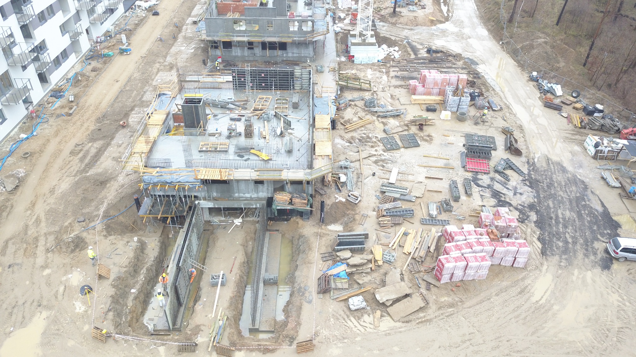 Widok z góry na plac budowy z fundamentami w wykopie, częściowo wylane ściany budynku w szalunkach, składowane materiały budowlane i robotników w żółtych kamizelkach.
