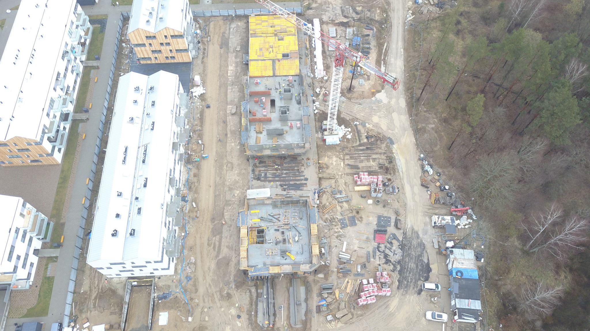 Widok z lotu ptaka na plac budowy z trzema budynkami w trakcie budowy, z widocznymi szalunkami, zbrojeniami i żurawiem wieżowym, otoczony nowymi budynkami mieszkalnymi i lasem.