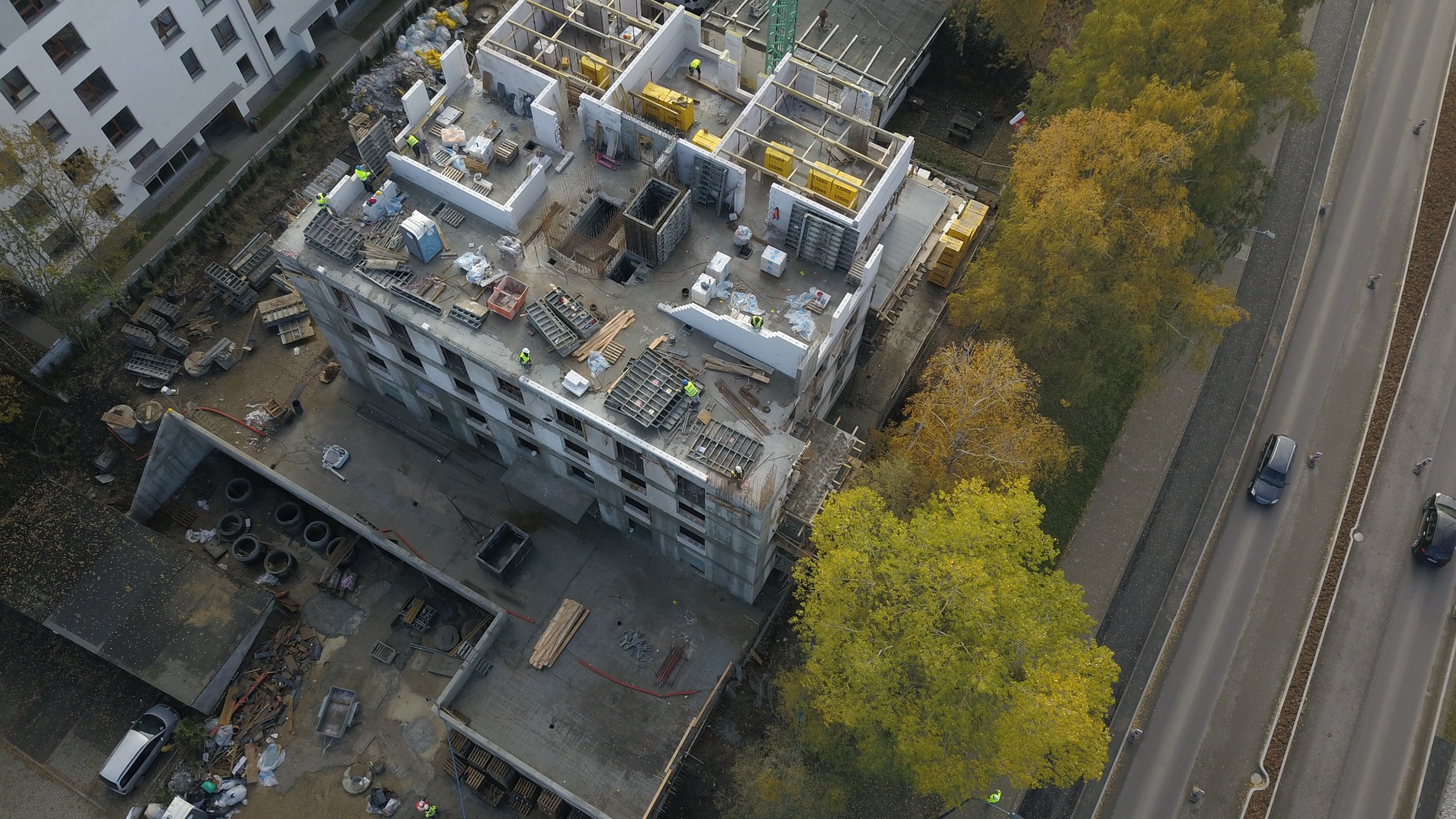 Widok z lotu ptaka na plac budowy budynku wielorodzinnego w trakcie realizacji, z widocznymi elementami konstrukcji żelbetowej, drewnianymi szalunkami i pracownikami w żółtych kamizelkach. Obok...