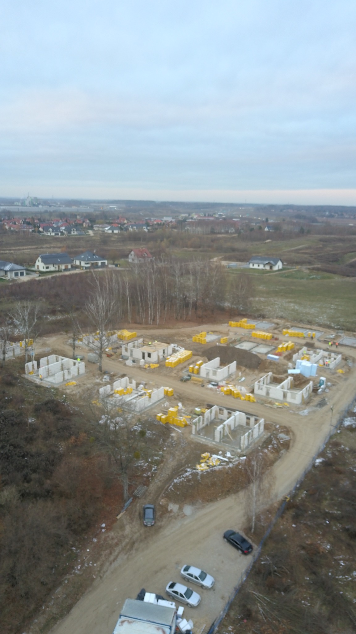 Widok z lotu ptaka na plac budowy z kilkoma domami w stanie surowym otwartym, materiały budowlane w żółtych opakowaniach, w tle zabudowa mieszkalna i pochmurne niebo.