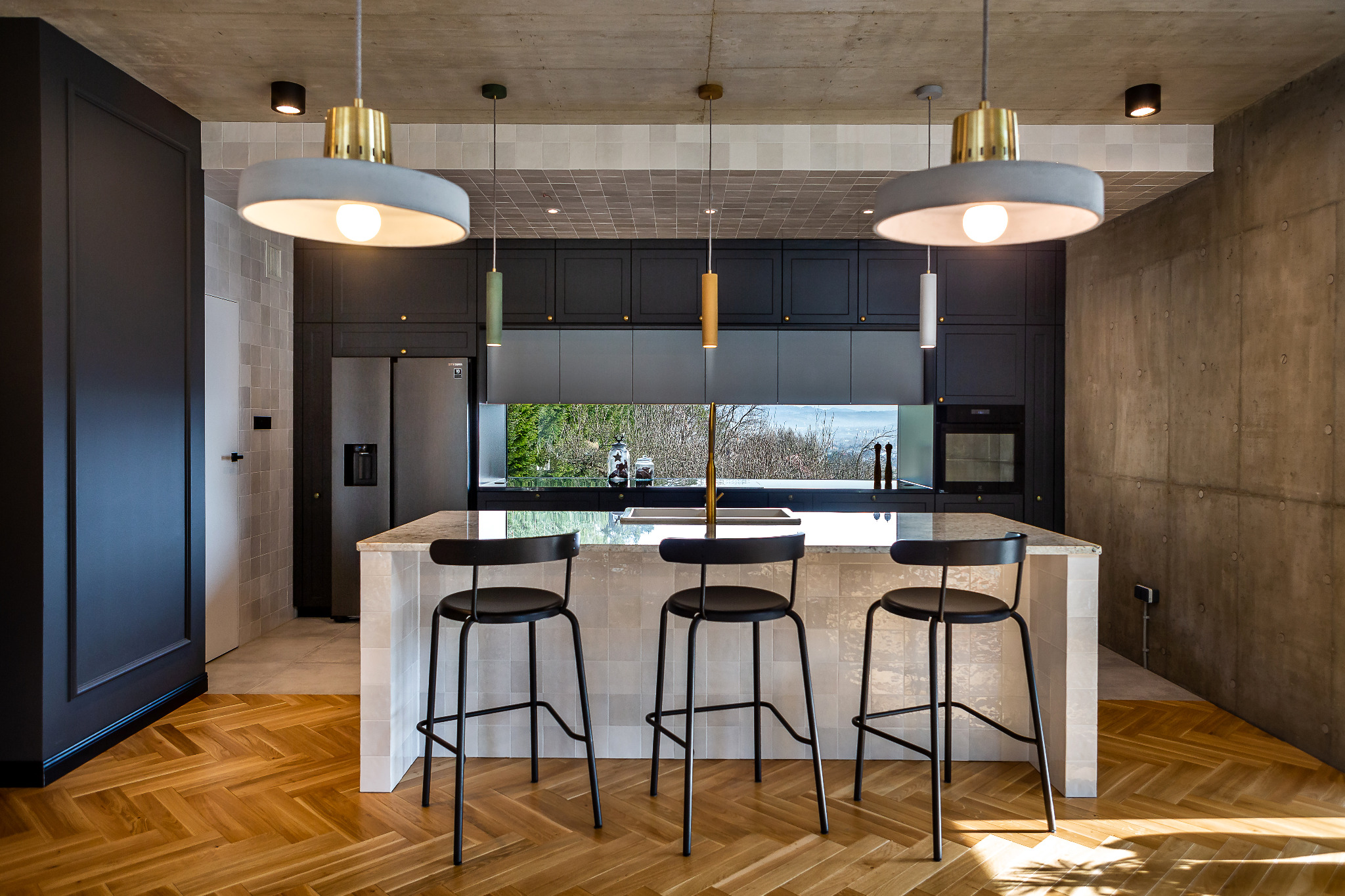 Nowoczesna kuchnia z wyspą, czarne szafki z subtelnymi złotymi detalami, betonowy sufit i ściana, parkiet w jodełkę, trzy czarne hokery przy wyspie, okno z widokiem na zieleń.