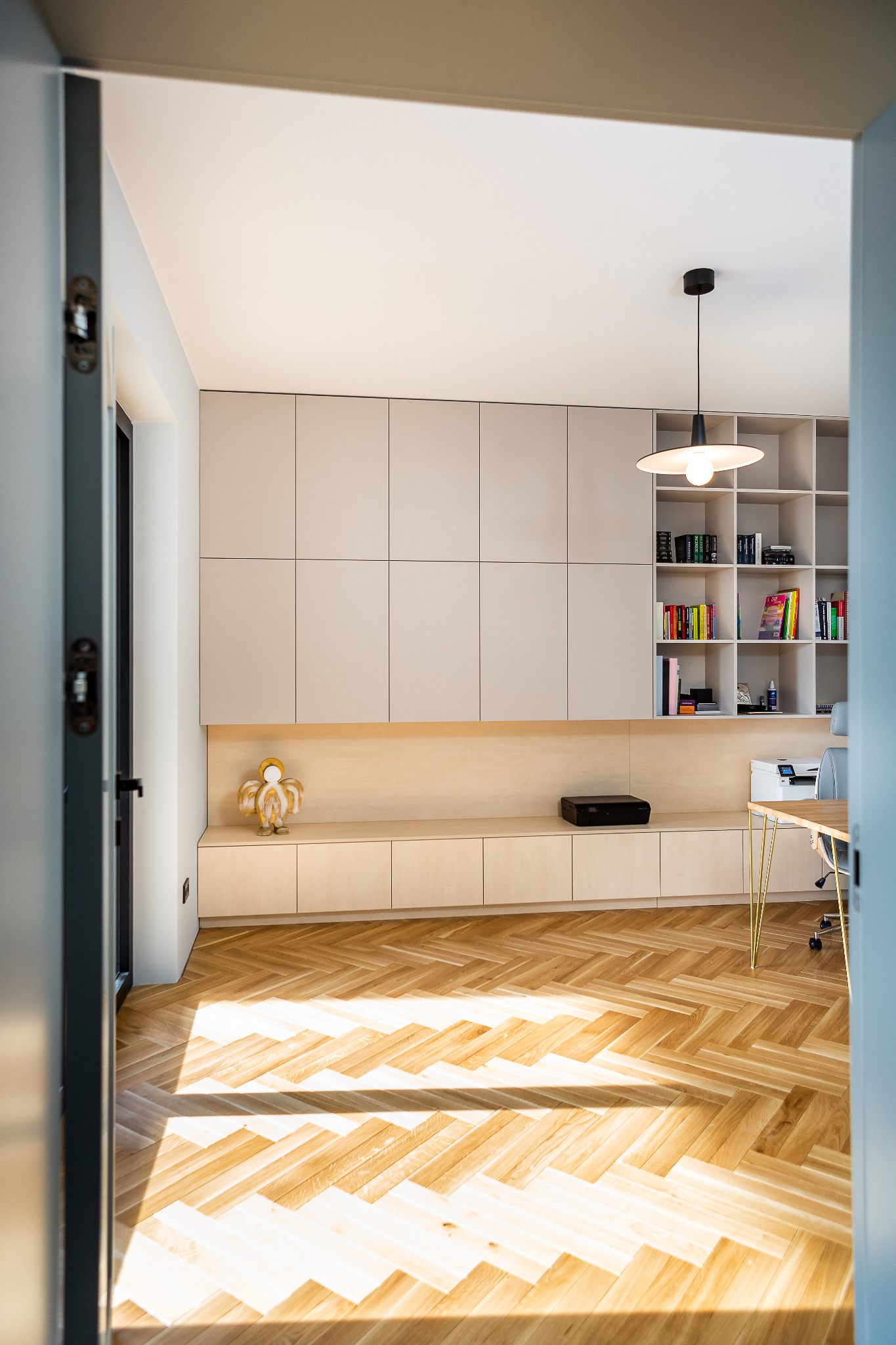 Nowoczesne biuro domowe z beżowymi szafkami na wymiar, drewnianym parkietem w jodełkę i złotymi nogami biurka, widziane zza uchylonych drzwi.