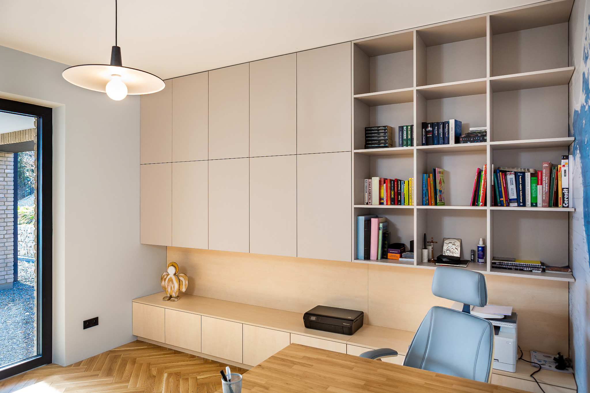 Jasne, nowoczesne biuro domowe z zabudową meblową: szafki, półki na książki, biurko z krzesłem, parkiet w jodełkę i widok na ogród przez okno.