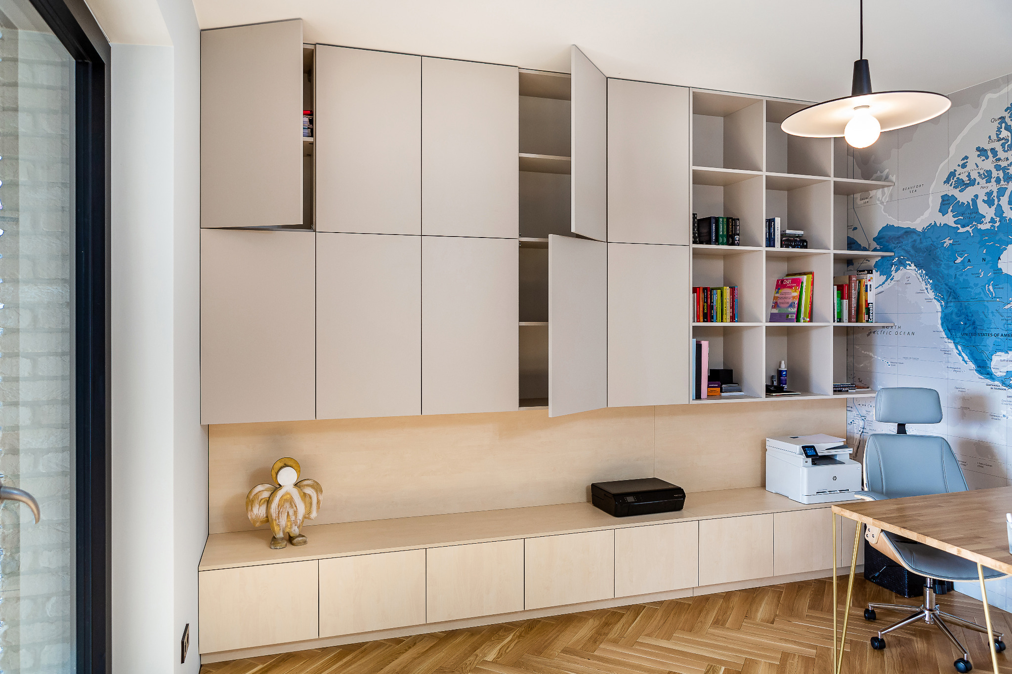 Zabudowa meblowa w jasnych odcieniach z szafkami, półkami i blatem, na którym stoi drukarka, obok biurko z krzesłem, a w tle mapa świata.