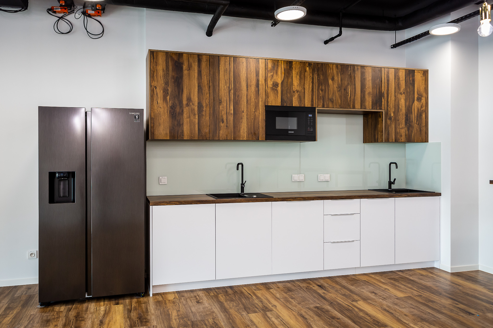 Nowoczesna kuchnia z białymi szafkami dolnymi, drewnianymi szafkami górnymi, czarnym zlewem i baterią, oraz dużą, ciemną lodówką side-by-side.