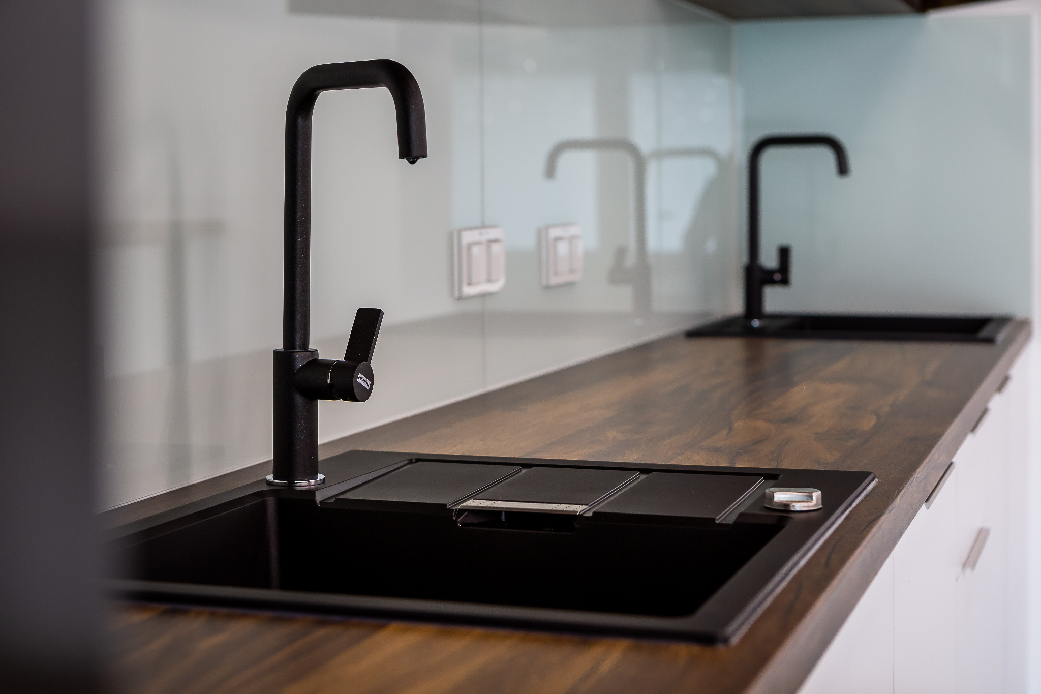 Nowoczesna kuchnia: czarny, kwadratowy zlew z matową, czarną armaturą Franke na blacie imitującym drewno, zintegrowany ociekacz, szklany panel ścienny w tle.