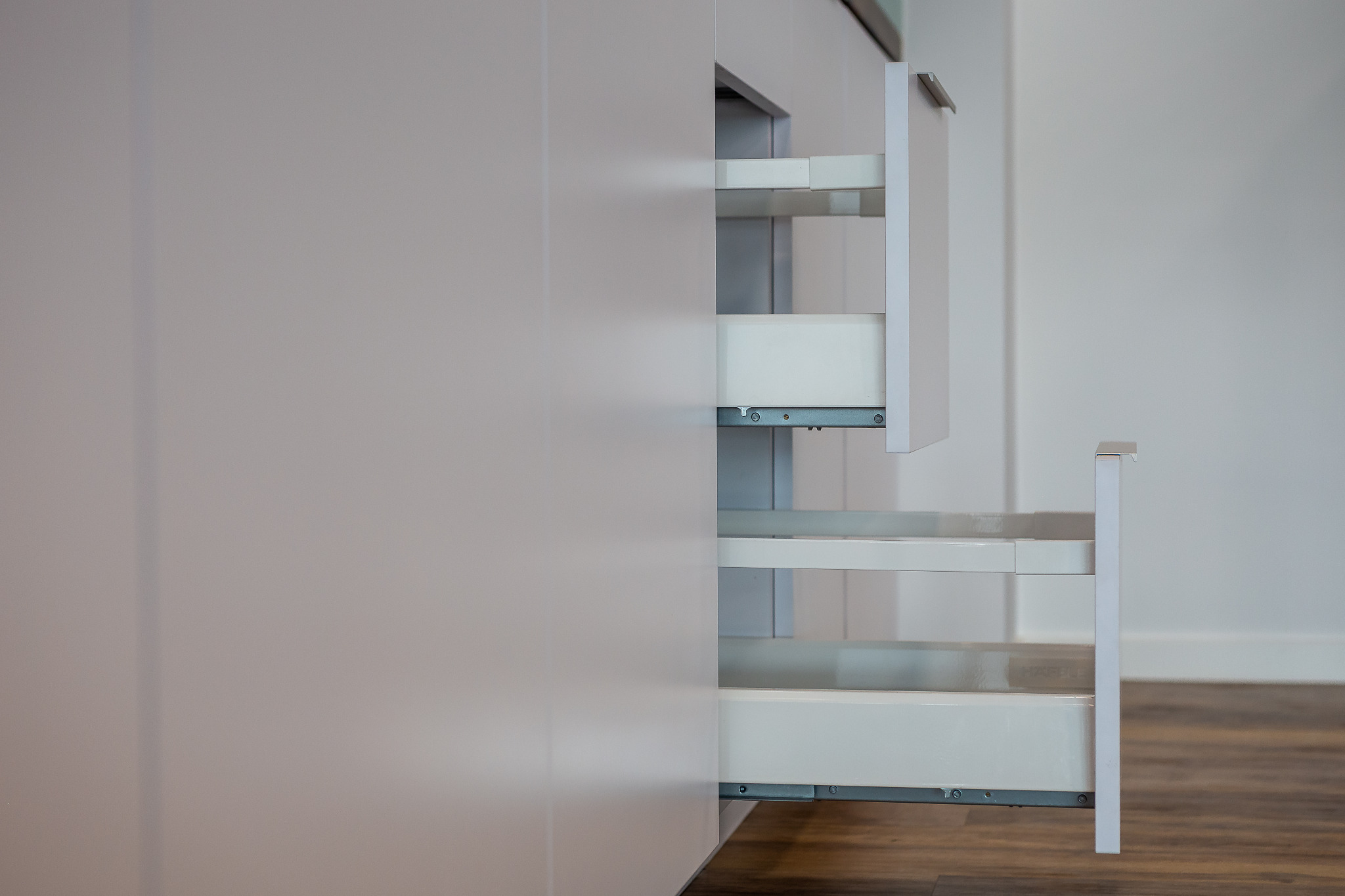 Wysuwane, białe półki cargo wewnątrz szafki kuchennej z matowymi, jasnoszarymi frontami, widoczne mocowania i prowadnice szuflad.