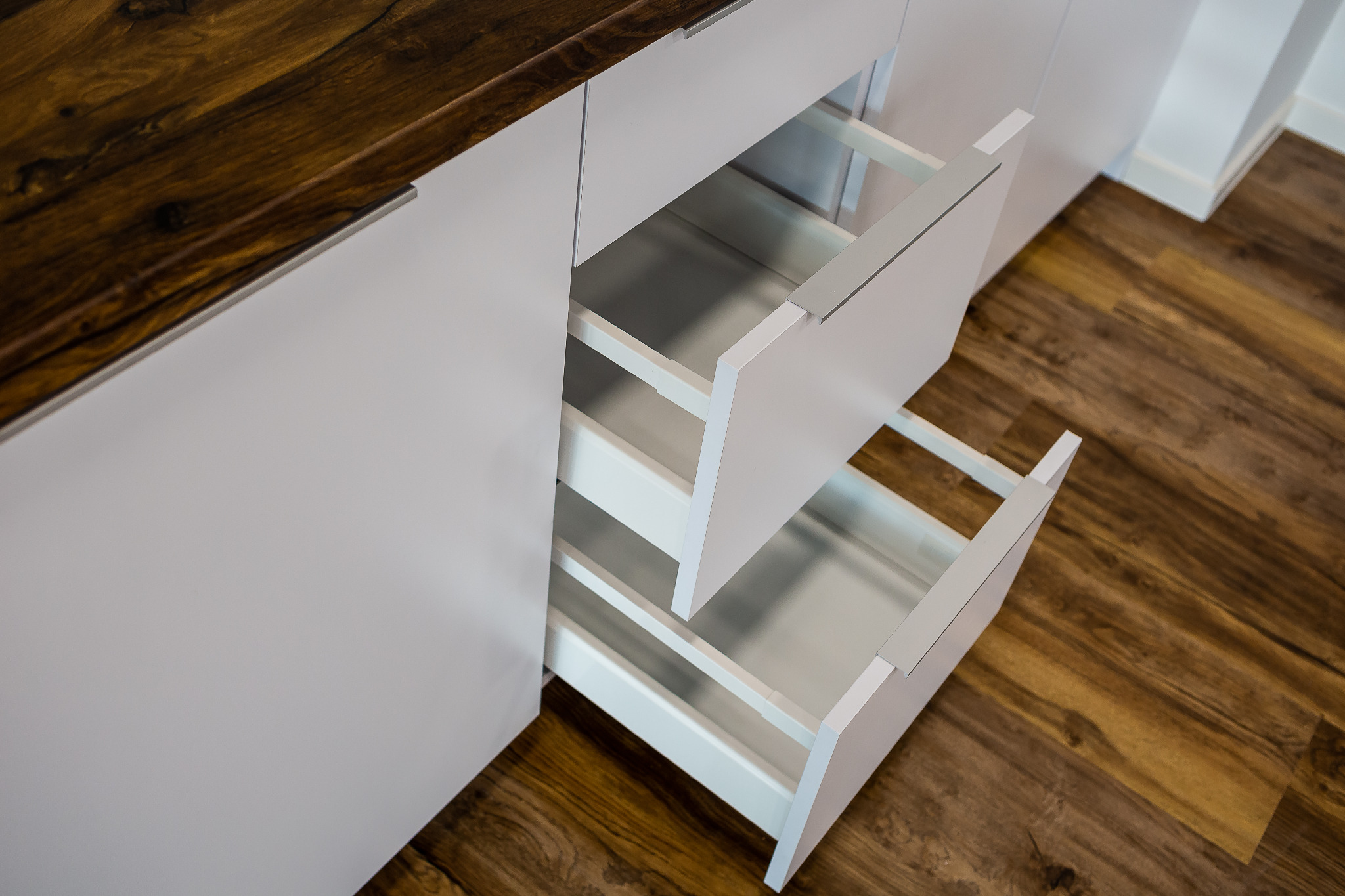 Białe szafki kuchenne z otwartymi szufladami, drewniany blat i podłoga w odcieniach brązu.