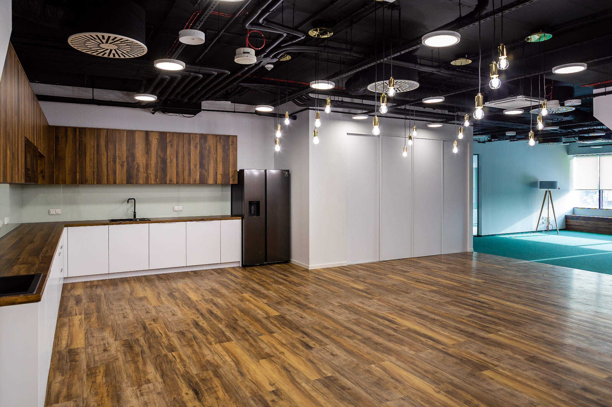 Nowoczesna kuchnia z drewnianymi szafkami górnymi, białymi dolnymi, czarnym zlewem i lodówką side-by-side w przestronnym biurze z drewnianą podłogą i industrialnym oświetleniem sufitowym.