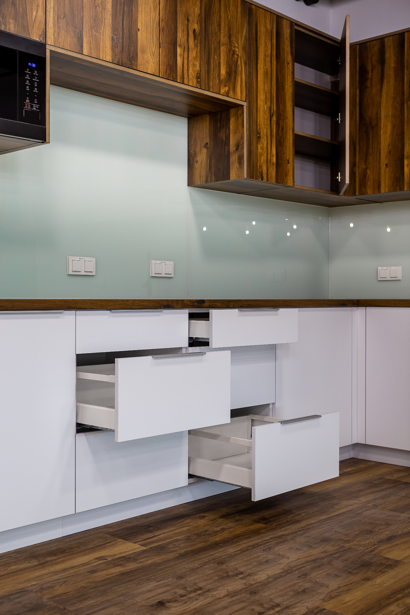 Nowoczesna kuchnia z białymi frontami i drewnianymi szafkami górnymi, otwarte szuflady z metalowymi uchwytami, szklany panel nad blatem, widoczny parkiet.