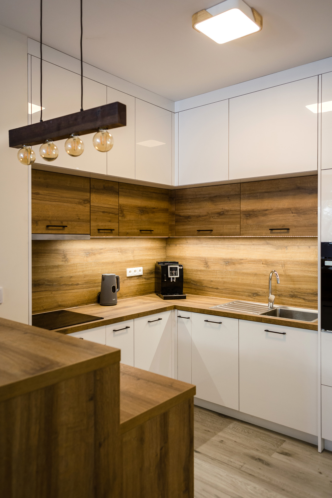 Nowoczesna kuchnia z białymi szafkami dolnymi i drewnianymi górnymi, oświetlenie LED pod szafkami, drewniany blat, ekspres do kawy i czajnik elektryczny, lampa wisząca z żarówkami dekoracyjnymi.